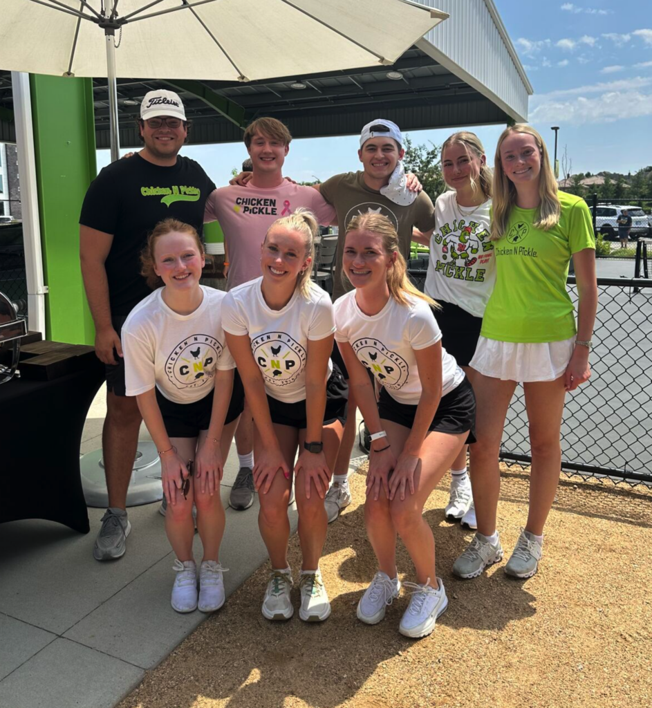 A group of eight young adults, dressed in sporty Chicken N Pickle shirts and shorts, pose and smile together outdoors under a large umbrella on a sunny day near a pickleball court.